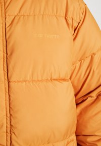 Oranje pufferjack met een gequilt ontwerp, een gladde textuur en subtiele branding in lichtgeel op de borst.