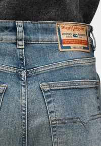 Modré džínsové nohavice s mierne opotrebovanou textúrou, výraznými zadnými vreckami a obdĺžnikovou štítkom s informáciami o značke prišitým na páse.