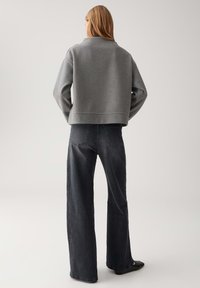 Donna in piedi di spalle, con indosso un pullover grigio a maniche lunghe e jeans neri a gamba larga, completati da scarpe nere a punta.