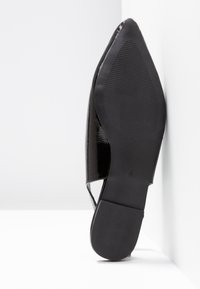 Black patent leather shoe featuring a pointed toe, open sides, and a textured rubber sole. The heel is flat and slim for a sleek design.