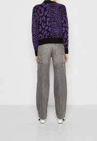 Pull violet et noir à motifs, associé à un jean gris à jambes larges avec des détails cloutés sur les poches et une coupe décontractée.