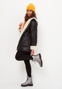 Manteau en fausse fourrure noire avec doublure crème, haut orange, leggings noirs et bottines argentées avec semelles noires. Le mannequin pose avec une jambe levée.