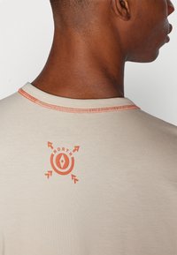 Camiseta beige de cuello redondo, con costuras naranjas. Un gráfico naranja con flechas y la palabra "NORTH" está impreso en la parte posterior.