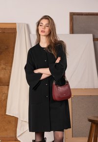 Czarny teksturowany płaszcz z rękawami o długości do łokcia w kształcie dzwonka, zapinany z przodu na guziki, zestawiony z małą bordową torebką z złotymi elementami.