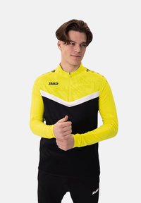 Maglia atletica a maniche lunghe di un vivace giallo con accenti neri; presenta un collo con zip, un motivo geometrico e il logo sul petto e sulla manica.
