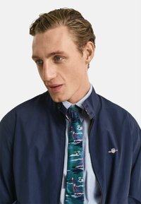 Giacca a vento blu navy con zip e logo, indossata sopra una camicia a righe blu e una cravatta con motivo di barche a vela multicolori, caratterizzata da texture vivaci.