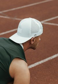 White cap with a flat brim, mesh sides for breathability, and a branded patch. Worn by a person in a green sleeveless top on a track surface.