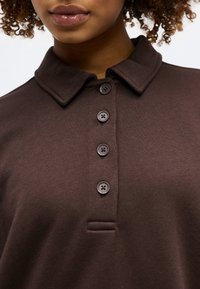 Γυναίκα που φοράει σκούρο καφέ πουκάμισο με γιακά, τέσσερα ορατά κουμπιά και υφασμάτινη υφή.