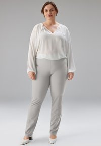 Weiße, fließende Bluse mit V-Ausschnitt und langen Ärmeln, kombiniert mit hellgrauen, taillierten Hosen und weißen, spitz zulaufenden High Heels. Glatte Stoffstrukturen.