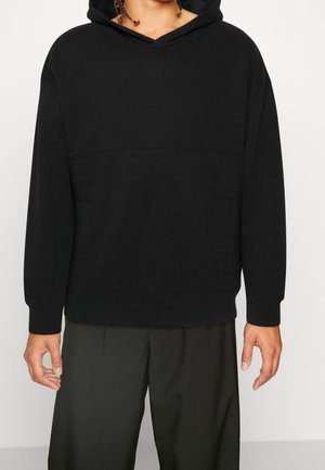 Person trägt einen schlichten schwarzen Kapuzenpullover mit langen Ärmeln und dunkelgrünen Hosen, steht vor einem weißen Hintergrund.