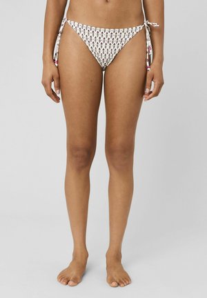 Femme portant un bas de bikini blanc à motifs floraux avec des liens sur les côtés, debout pieds nus devant un fond gris uni.