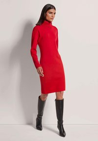 Rotes Strickkleid mit hohem Kragen, gerippten Schulterdetails und taillierter Silhouette, kombiniert mit schwarzen knielangen Stiefeln.