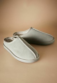 Chaussons en suède gris à enfiler, avec un intérieur doux et doublé, et des coutures contrastantes le long des bords, sur fond neutre.
