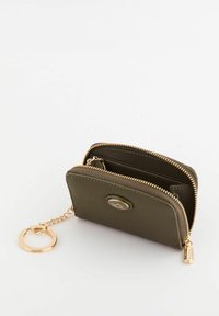 Cartera con cremallera en verde oliva, con un lazo para llavero en tono dorado, acabado texturizado y un logo decorativo en la parte frontal. Se abre para revelar dos compartimentos.