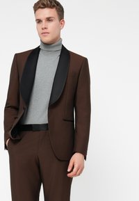 Tânăr îmbrăcat într-un costum maro cu reverne negre peste un pulover gri cu gât înalt, stând în fața unui fundal alb.