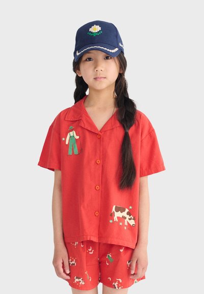 Enfant avec de longues tresses foncées portant une casquette bleu marine et un ensemble rouge deux pièces avec un motif de fermier et de vache sur le t-shirt et le short.