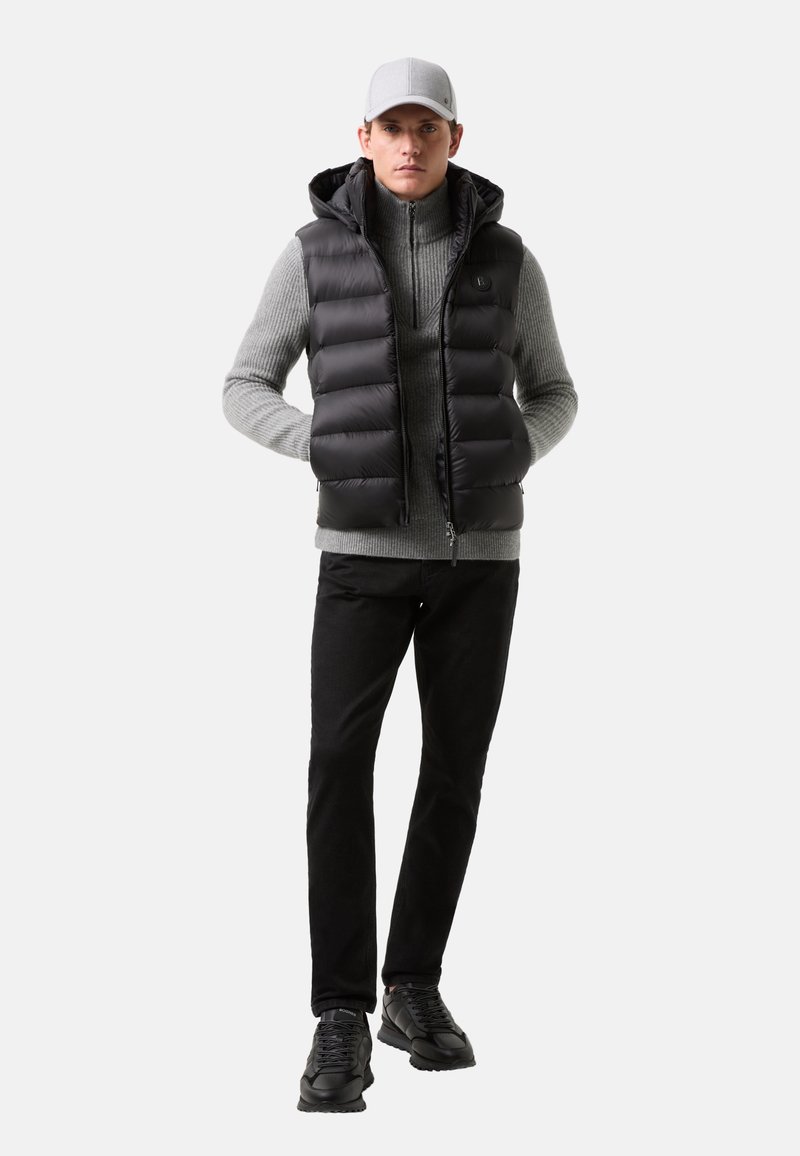 Schwarze Pufferweste mit Kapuze über einem grauen gestreiften Rollkragenpullover, kombiniert mit schwarzen Hosen und schwarzen Sneakers. Das Model trägt eine graue Mütze.