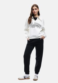 Hvid sweatshirt med "New Jersey Athletic" tryk, parret med sorte joggers. Modellen bærer hvide sneakers med sorte og grønne accenter.