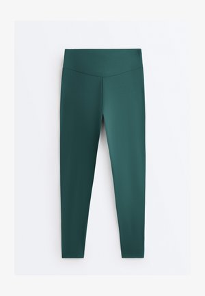 Donkergroene leggings met een hoge taille, een gladde, naadloze afwerking en nauwsluitende pijpen tot aan de enkels, plat neergelegd op een witte achtergrond.