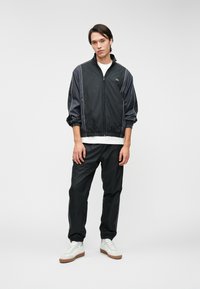 Μαύρο αδιάβροχο με γκρι λεπτομέρειες, φερμουάρ στο κλείσιμο και ελαστικές μανσέτες, συνδυασμένο με μαύρο παντελόνι και λευκά αθλητικά παπούτσια με καφέ σόλα.