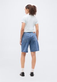 Γυναίκα με κατσαρά μαλλιά, ντυμένη με λευκό πόλο πουκάμισο, μπλε τζιν σορτς, λευκές κάλτσες και μαύρα παπούτσια, στέκεται με την πλάτη προς το μέρος μας σε απλό φόντο.