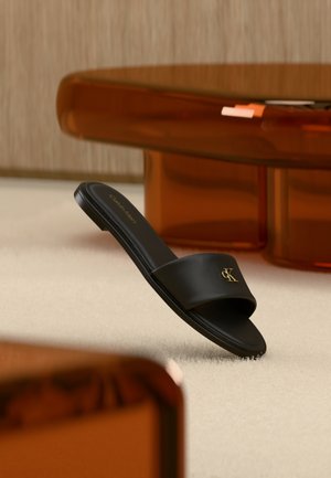 Sort Calvin Klein slip-in sandal med gyldent logo, hvilende på et beige tæppe, i nærheden af et gennemsigtigt ravfarvet bord med stabel af bøger.