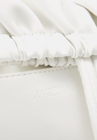 Valkoinen nahkalaukku, jossa on ryppynyt rakenne, sileä pinta ja kohokuvioitu "MARCCAIN"-logo sivussa.