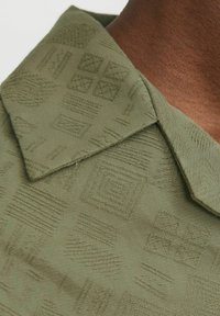 Olijfgroene overhemd met kraag, voorzien van een textuur van geometrische patronen en een gladde afwerking. Zichtbare stiksels langs de randen van de kraag.