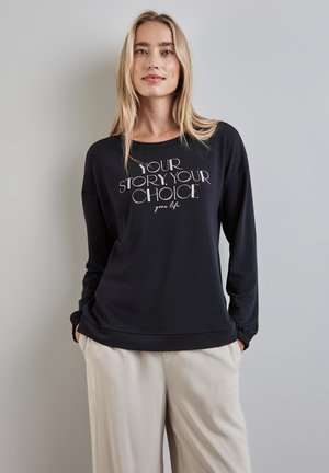 Schwarzer Langarm-Sweatshirt mit weißem Text "DEINE GESCHICHTE, DEINE WAHL", in entspannter Passform und mit gerippten Bündchen, kombiniert mit beigen Hosen.