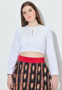Top bianco corto con dettagli in pizzo sul décolleté, scollatura rotonda e maniche lunghe; abbinato a una gonna nera con motivo floreale e fascia rossa in vita.