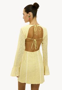 Vestido amarillo de encaje con mangas largas acampanadas y espalda descubierta, con un cierre de lazo. Patrón geométrico con acentos blancos en el frente.