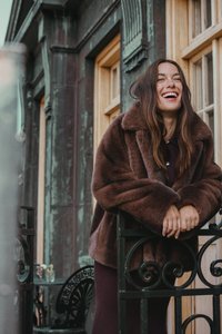 Kobieta w brązowym futrze opiera się o czarną żelazną bramę, uśmiechając się, z oknami zabytkowego budynku w tle.