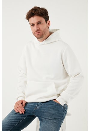 Biela mikina s kapucňou z mäkkého materiálu s predným vreckom a nastaviteľnou šnúrkou na kapucni, doplnená modrými džínsami.