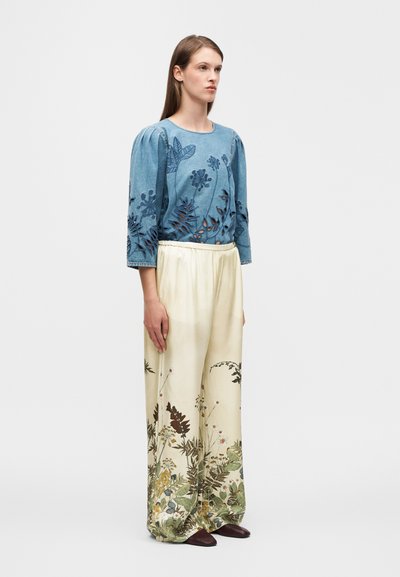 Τζιν μπλούζα με λουλουδάτα κοψίματα, μπλε, συνδυασμένη με κρεμ παντελόνια φαρδιά στο πόδι με βοτανικό σχέδιο στο τελείωμα. Καφέ παπούτσια.