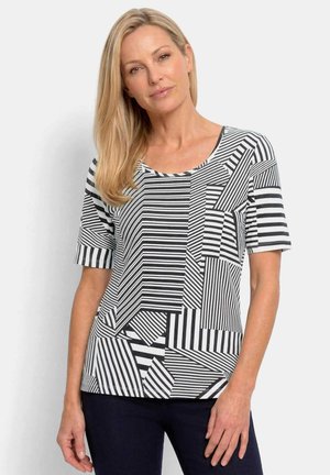 Femme blonde portant un haut à manches courtes noir et blanc avec des motifs rayés mélangés, debout devant un fond clair uni.
