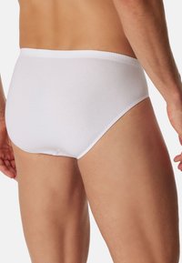 Uomo che indossa slip bianchi semplici, mostrato di spalle, evidenziando la vestibilità e la trama del tessuto su uno sfondo uniforme.