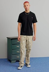 T-shirt nera a maniche corte con tasca, pantaloni beige a fantasia e sneakers neri. Persona in piedi accanto a un cassettone verde scuro.