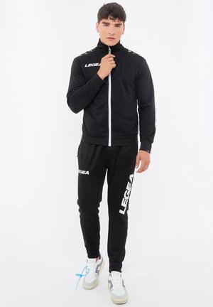 Man draagt een zwart Legea trainingspak met witte rits en logo's, hij stelt de kraag van zijn jas bij en staat voor een witte achtergrond.