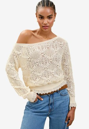 Maglione in pizzo color crema con scollo a barca, caratterizzato da motivi intricati e polsini a coste, indossato sopra jeans high-waisted di un delicato blu chiaro.