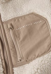 Veste en polaire avec une texture crème et une poche beige dotée d'une fermeture éclair et du texte imprimé "AVENTURES À VENIR". Détails brodés le long des bords.