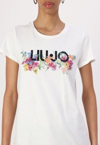 T-shirt en coton blanc avec un imprimé floral comprenant des fleurs colorées et des lettres "LIU.JO" en noir, à manches courtes et col rond.
