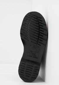 Black rubber shoe sole with a textured pattern, featuring deep grooves for traction and a central logo embossing.