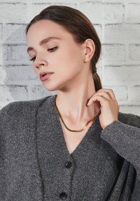 Grijze gebreide cardigan met zwarte knopen, gedragen over een gouden ketting. De achtergrond heeft een getextureerde witte bakstenen muur.
