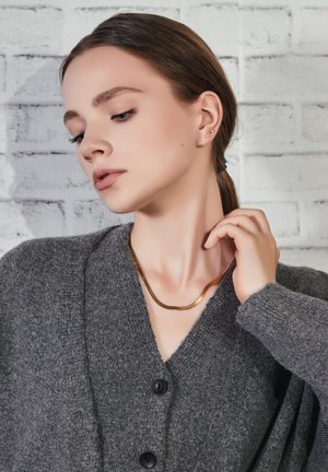 Harmaa neulottu neuletakki mustilla napilla, kerrostettuna kullanvärisen kaulaketjun päälle. Taustalla on teksturoitu valkoinen tiiliseinä.