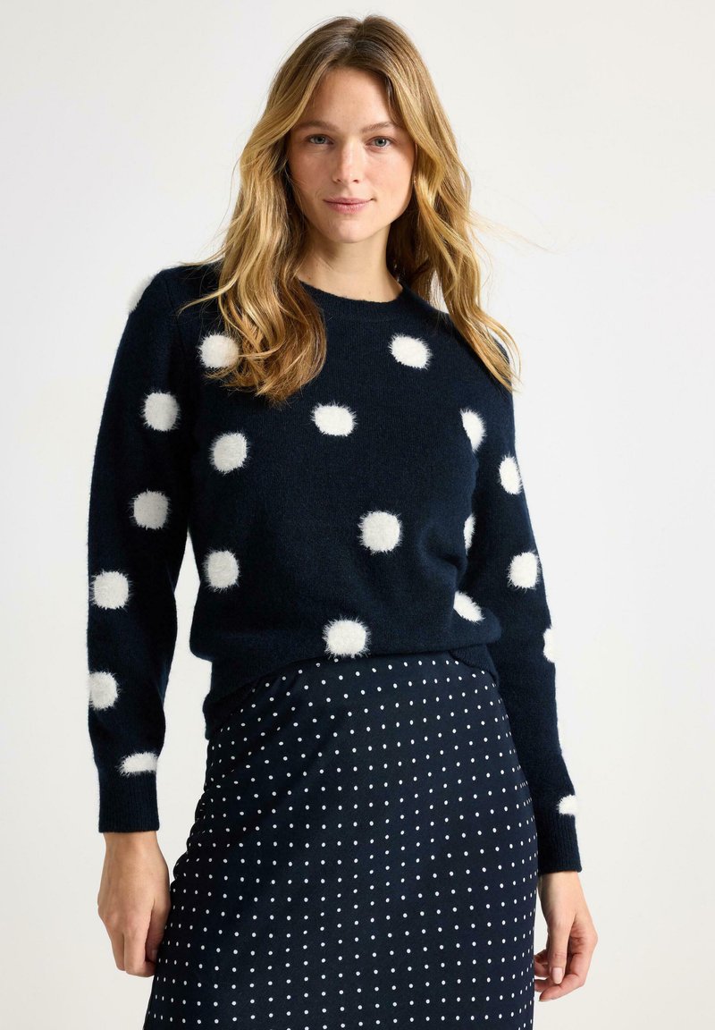 Maglione blu navy con pois bianchi soffici, scollo rotondo e maniche lunghe; abbinato a una gonna scura con pois bianchi più piccoli.