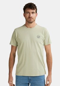 Hellgrünes Baumwoll-T-Shirt mit Rundhalsausschnitt und kurzen Ärmeln, das ein kleines rundes Logo auf der linken Brust trägt. Schlichte, glatte Textur.