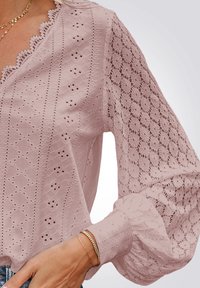 Blouse rose clair avec des détails en dentelle, encolure festonnée et manches bouffantes. Présente des motifs ajourés et un tissu texturé avec des poignets superposés.