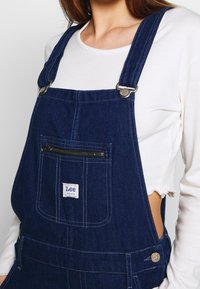 Ciemnoniebieskie ogrodniczki denimowe z regulowanymi ramiączkami, przednią kieszenią i detalem z kieszenią na zamek. Zawierają naszywkę z logo Lee na przodzie.