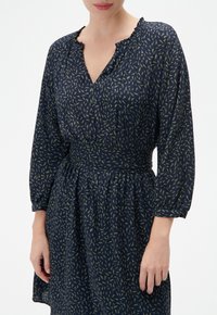 Marineblaues Kleid mit grünem Blattdruck, mit einem Rüschenausschnitt, drei Viertel-Ärmeln und einer taillierten Taille, die zu einem schwingenden Rock übergeht.