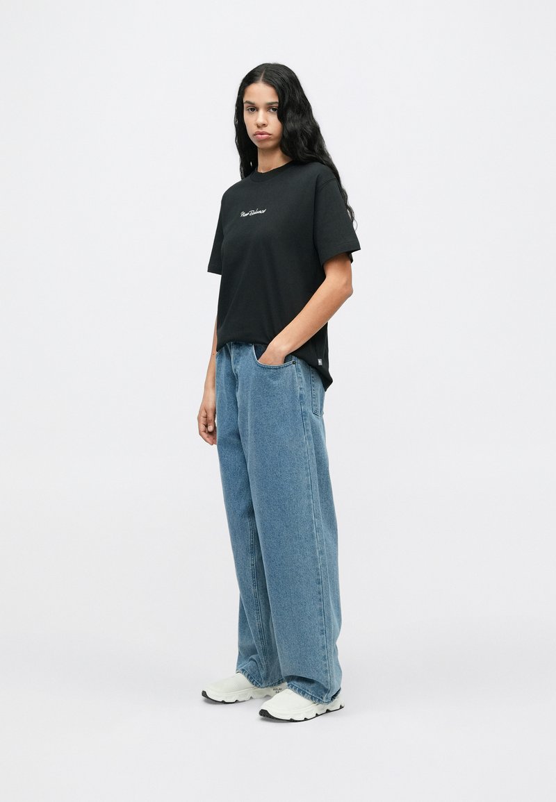 T-shirt nera in cotone con maniche corte e ricamo sul petto, abbinata a jeans in denim azzurri, a vestibilità rilassata, e sneaker bianche.